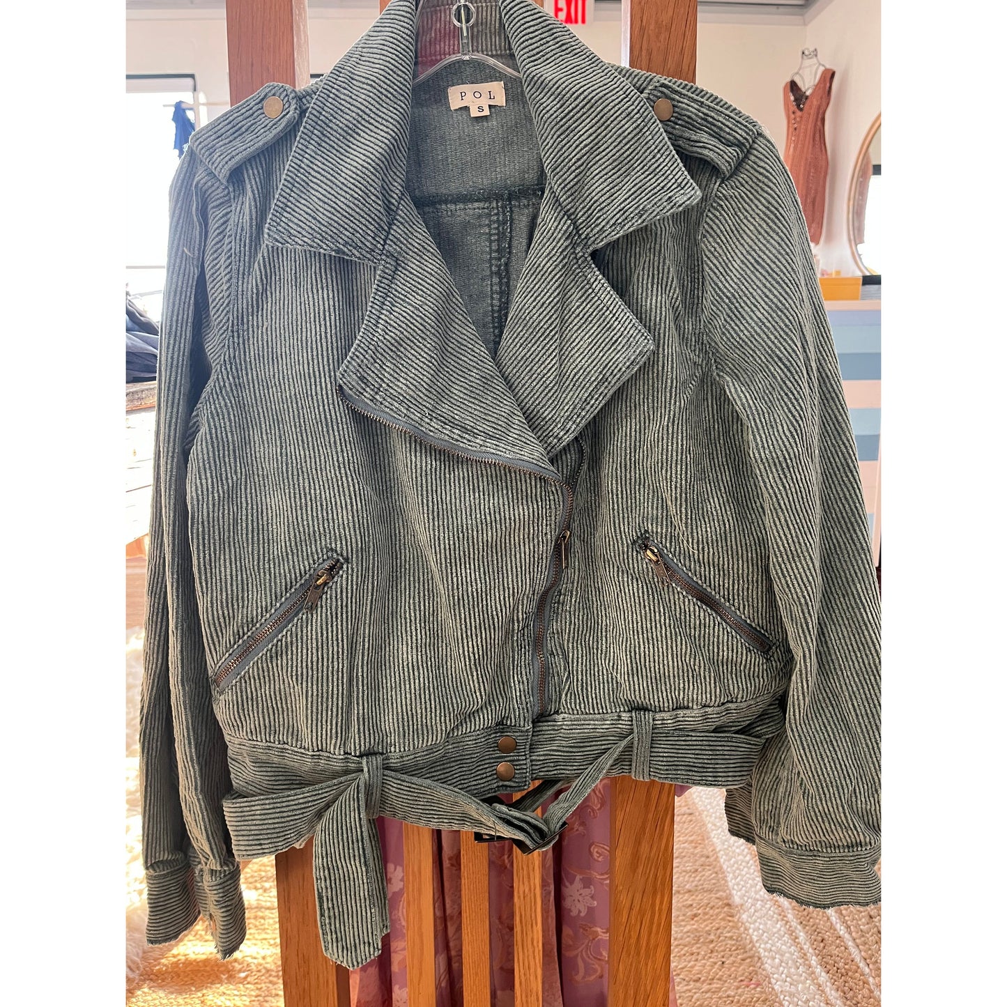 Hunter Green, Corduroy Jacket with diagonal zipper detail going from the left hip to the right shoulder. The bottom of the jacket features two stacked silver buttons on either side of the front opening below the zipper. Belt loops attached around the base hem with a tie belt. There are 2 diagonal pockets on either side of the jacket. Large double flaps are featured on the front, 100% Cotton. Available in sizes Small - Large