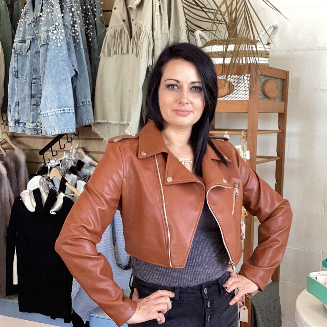 Tan faux leather cropped jacket with zipper in the front. There are buttoned down shoulder straps on the tops of the shoulders. Double flaps with button detail on the tips of the flaps. There is one small zippered pocket on the left side around the chest area. chest   Available in sizes Small through Large. Faux Leather.